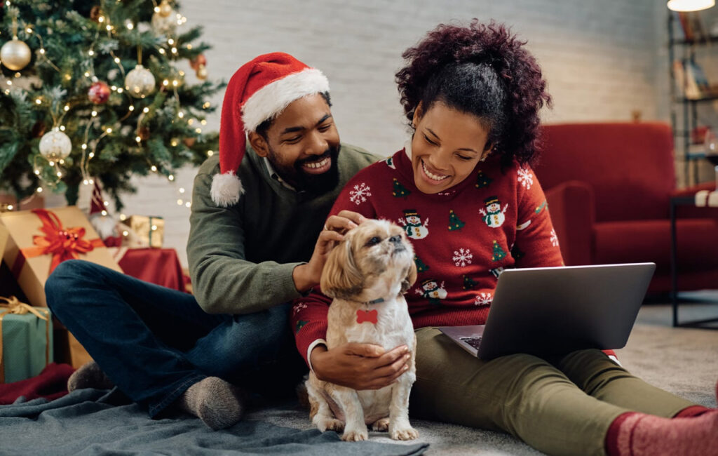 Couple in Christmas sweaters smiling at a laptop with a dog in front of a decorated tree.